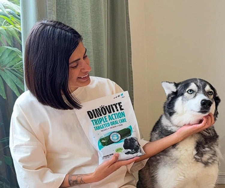 A woman sits beside a husky and holds up a bag of Dinovite Triple Action Targeted Oral Care chews.